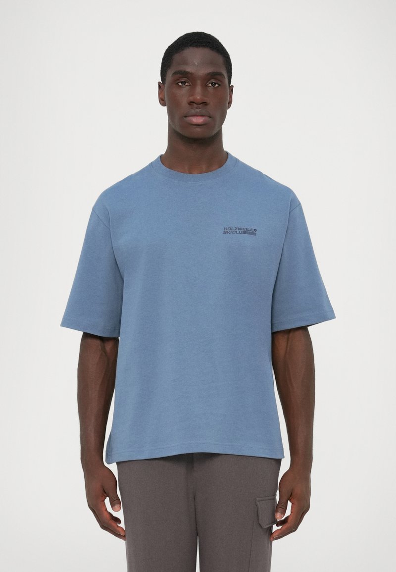 T-shirt de algodão azul com mangas curtas, decote redondo e um logótipo impresso em fonte escura no lado esquerdo. Usada com calças cinzentas de corte solto.