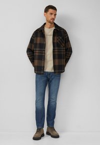 Zwarte en bruine geruite shirtjack, beige crewneck t-shirt, blauwe spijkerbroek en bruine veterschoenen. Gedurfde ruitpatroon en detail van het shirtzakje.