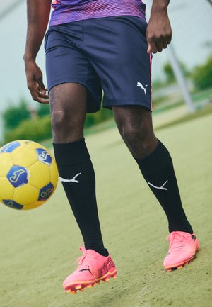 Puma fodboldstøvler i livlig pink med struktureret detaljering, sorte sokker med hvidt logo og marineblå shorts på en grøn bane.