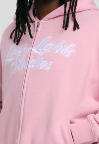 Sweat à capuche zippé rose en tissu doux, avec le texte brodé « Low Lights Studios » en blanc. Comprend une poche avant et une capuche.