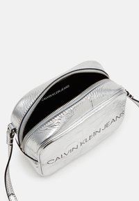 Stříbrná metalická crossbody kabelka s hladkým povrchem, vybavená dvojitým zipem, černým interiérem a potištěným logem "CALVIN KLEIN JEANS".