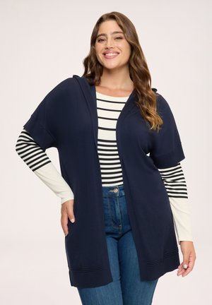 Kvinde smilende, iført en marineblå kortærmet åben cardigan over en sort og hvid stribet langærmet top med blå jeans.