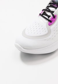 Chaussure de sport blanche avec une tige en mesh texturé, des accents argentés, des œillets violets et des lacets noirs, dotée d'un bout arrondi et d'une semelle coussinée.