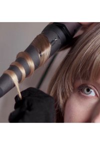 Krultang met een zwart handvat, verwarmde cilinder die een lok lichtbruin haar vasthoudt. Close-up van een hand in een zwarte handschoen en het oog van een persoon.