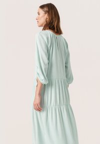 Mintgrøn, lagdelt kjole med struktureret stof, trekvartærmer og en nøglehulldetalje bagpå. Enkel og flydende design.