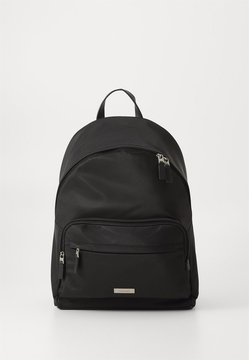 Mochila negra de nylon con parte superior redondeada, dos compartimentos con cremallera y un asa de tela. Herrajes plateados y una placa de logo en el frente.