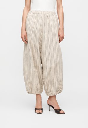 Kvinde iført beige vidde bukser med lodrette tynde striber og elastiske kanter, kombineret med sorte åbne hælede muleposer.