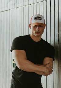 Čierna čiapka s bielou prednou časťou a oranžovým šiltom; čierne priliehavé tričko. Subjekt má svetlú pleť a je umiestnený na pozadí z dreva.