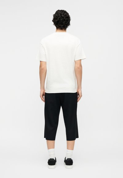 Homme aux cheveux bouclés et foncés, vu de dos, portant un t-shirt blanc uni à manches courtes, un pantalon noir court, des chaussettes blanches et des baskets noires.
