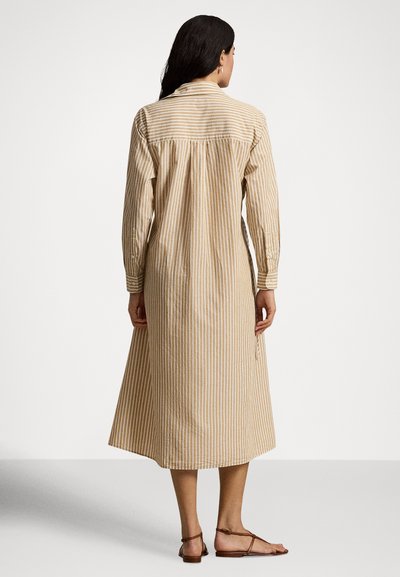 Femme debout, vue de dos, portant une robe midi à manches longues à rayures verticales beige et blanche, avec des sandales plates marron clair.