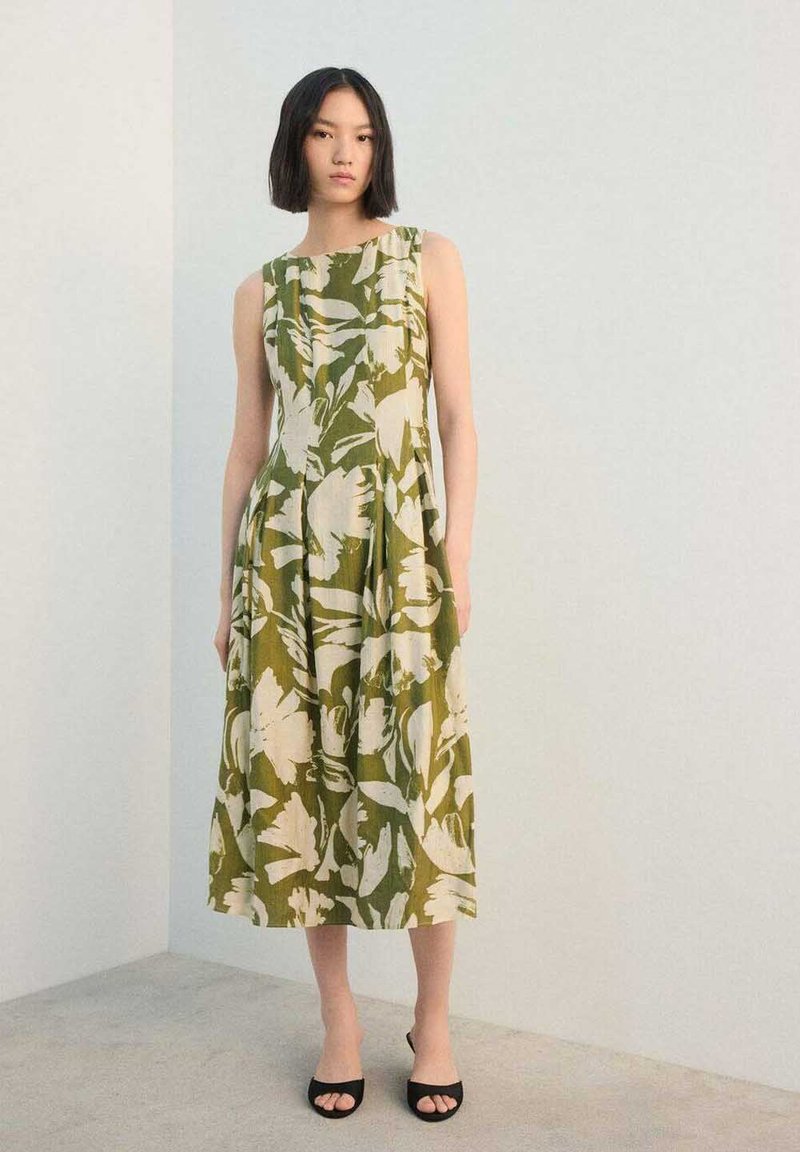 Robe à imprimé floral vert avec encolure bateau, design sans manches et jupe plissée. Chaussures noires à bouts ouverts complètent la tenue.