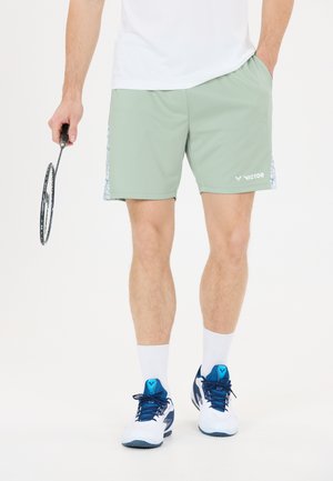 Mand iført lysegrønne Victor sportsshorts, hvide sokker, blå og hvide sneakers, holder en badmintonketcher, den ene hånd i lommen.