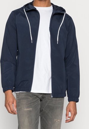 Homme portant une veste à capuche zippée bleu marine avec cordons blancs, un t-shirt blanc et un jean gris délavé légèrement usé.