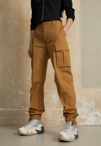 Cargo byxor i senapsgul tyg med cargo-fickor, tapered anklar och en texturerad finish, paret med chunky sneakers i ljusgrått och svart.