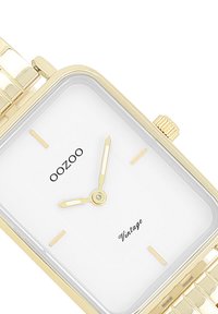 Montre rectangulaire dorée avec un cadran blanc, des index horaires minimalistes et un bracelet doré assorti. Comprend une couronne texturée et le texte "OOZOO Vintage".