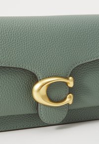 Gros plan d’un sac en cuir vert texturé avec une boucle métallique dorée courbée en forme de lettre C sur le rabat avant.