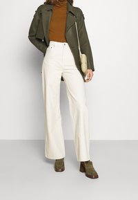 Gabardina verde oliva, suéter de cuello alto marrón, pantalones de pierna ancha color crema y zapatos de tacón verde oliva. Pequeño bolso bandolera beige.