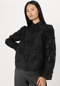 Blusa preta transparente com detalhes bordados de flores e lantejoulas. Apresenta colarinho e mangas compridas. Combinada com calças cinza listradas.