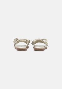Sandali piatti beige con ampi fiocchi laterali, design aperto sulla punta e suole color ocra, mostrati da dietro su uno sfondo bianco.