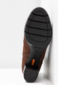 Chaussure en daim marron avec une tige texturée et une semelle en caoutchouc noire présentant des rainures horizontales et un accent de logo orange au talon.