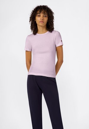 Kobieta o kręconych włosach, ubrana w jasnofioletową koszulkę z krótkim rękawem i ciemne spodnie, stoi z rękami za plecami na białym tle.