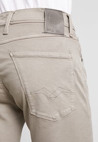 Jeans de mezclilla beige con un bolsillo trasero que presenta un diseño de costura en zigzag y una etiqueta de cuero de marca en la cinturilla. Textura suave.