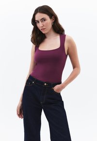 Bodysuit burdeos sin mangas con escote cuadrado, combinado con pantalones de mezclilla oscuros de cintura alta que presentan herrajes dorados y bolsillos laterales.