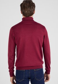 Een man staat met zijn rug naar de kijker, gekleed in een bordeauxrode coltrui en blauwe jeans, tegen een effen lichte achtergrond.