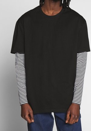 Person iført sort kortærmet t-shirt over en sort-hvid stribet langærmet skjorte, kombineret med blå jeans, mod en ensfarvet baggrund.