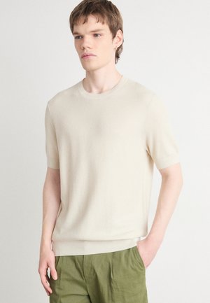 Maglione lavorato a maglia beige a maniche corte, con texture a coste, colletto rotondo e vestibilità rilassata. Abbinato a pantaloni verdi di taglio largo.
