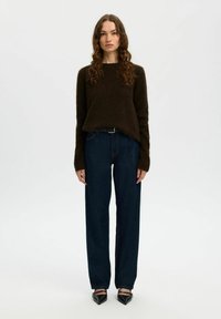 Donna in piedi di fronte, con un maglione marrone scuro, jeans blu scuro con una cintura nera e scarpe a punta nere.