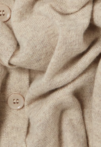 Cardigan en tricot beige avec une texture douce, présentant des boutons ronds de couleur claire et un tissu drapé créant une apparence décontractée et superposée.