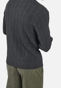 Tummanharmaa cable-knit-neule, jossa on ribattu hihansuut ja helma, yhdistettynä vaaleanvihreisiin housuihin. Tekstuuriltaan paksu ja kudottu.