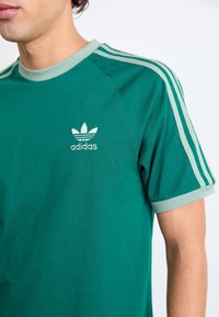 Grünes Baumwoll-T-Shirt mit Rundhalsausschnitt, kontrastierenden hellgrünen Akzenten an den Ärmeln und drei Streifen an den Schultern. Mit einem gestickten Logo versehen.