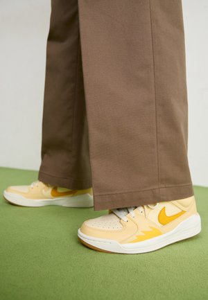 Osoba ubrana w brązowe spodnie o szerokich nogawkach i żółte buty sportowe Nike, stojąca na zielonej powierzchni na neutralnym tle.