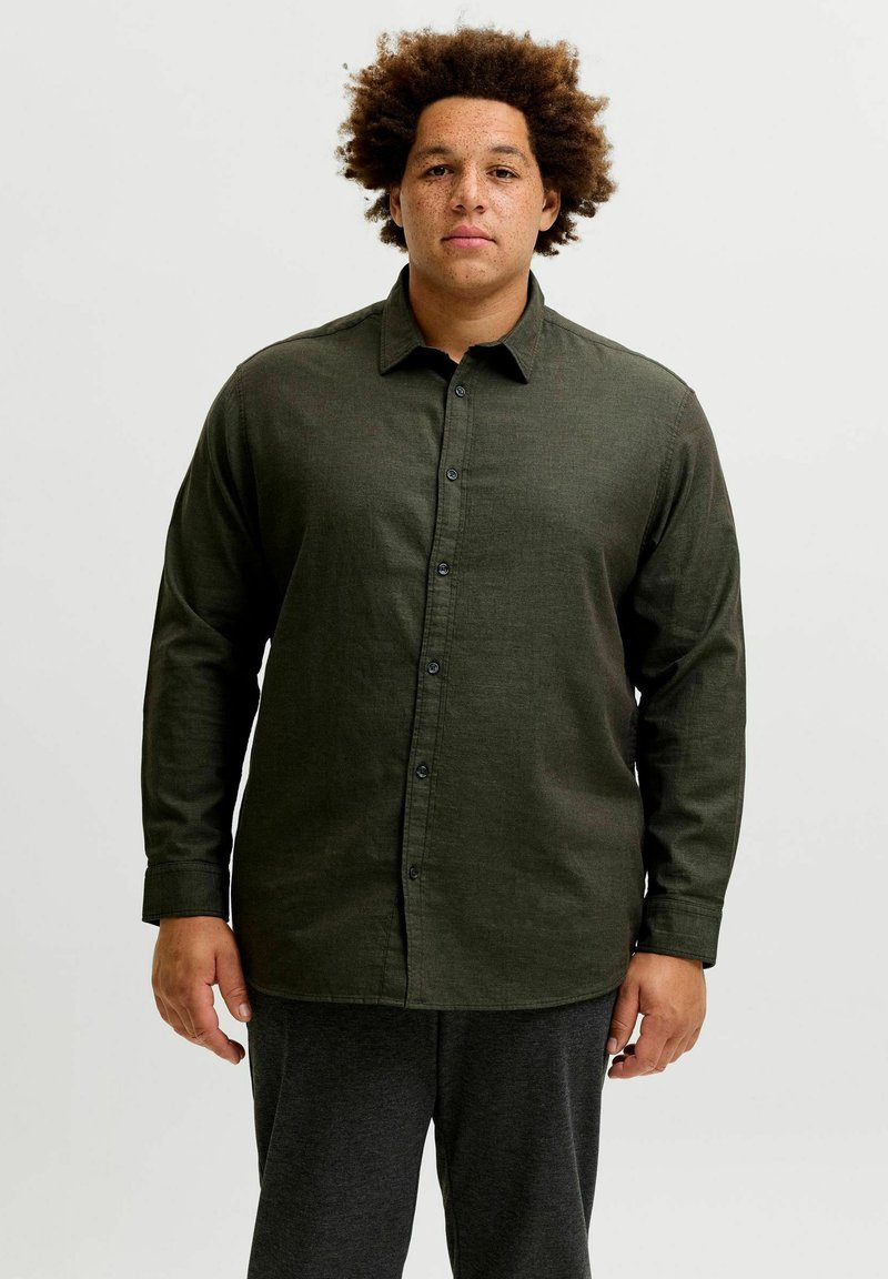 Άνδρας με σγουρά μαλλιά και φακίδες, που φοράει σκούρο πράσινο πουκάμισο με κουμπιά και σκούρο παντελόνι, στέκεται μπροστά σε απλό λευκό φόντο.