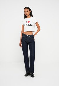 Femeie în picioare pe un fundal alb, purtând un tricou alb scurt cu inscripția "I ♥ PARIS", blugi negri și mocasini negri, pozând cu o mână pe șold.