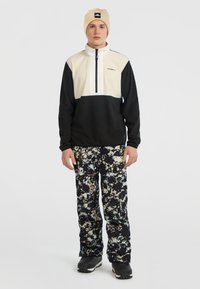 Pullover jack met een halve rits in crème en zwart; zwart-wit gespetterde broek; zwarte schoenen; beige muts met logo.