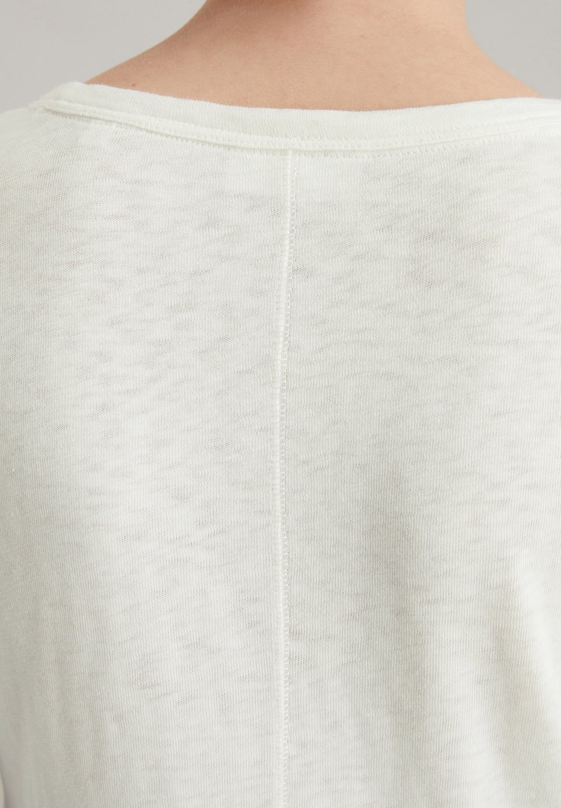 Jasny sweter dzianinowy o miękkiej teksturze, z szerokim dekoltem i centralnym szwem biegnącym wzdłuż pleców.