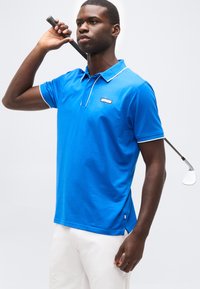 Homme en polo bleu et pantalon blanc tenant un club de golf derrière ses épaules, debout devant un fond gris uni.