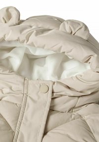 Beige vadderad jacka med ett mjukt vitt fleecefoder, som har en rund huva och små öronliknande detaljer, stängd med en tryckknapp.