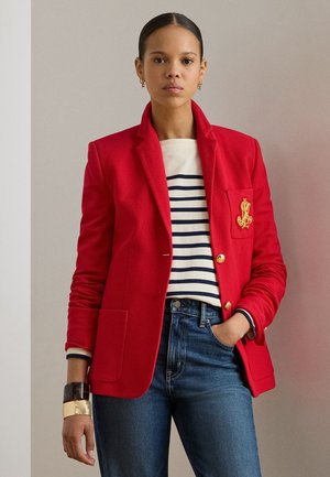 Blazer roșu din țesătură texturată, cu un emblemă aurie pe buzunar, purtat cu o cămașă în dungi și blugi din denim închis la culoare.