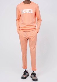 Camisa de manga larga color melocotón con el logo "HUGO", combinada con pantalones ajustados a juego y zapatillas multicolores. Diseño sencillo, textura suave.