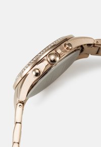 Montre en or rose avec un boîtier rond, ornée de petits cristaux le long de la lunette, dotée de boutons de commande et d'un bracelet en maillons métalliques.