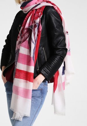 Femme portant un jean bleu, une veste en cuir noire et une grande écharpe à motifs floraux rose, rouge et blanc enroulée autour du cou.