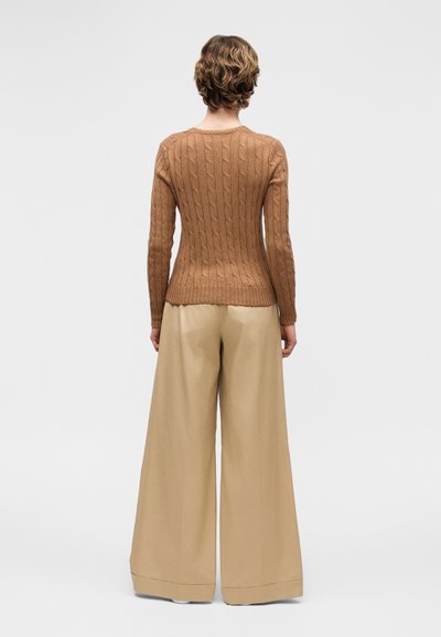 Άτομο με κοντά κυματιστά μαλλιά που φοράει καφέ πλεκτό πουλόβερ με σχέδιο καλώδιο και φαρδιά μπεζ παντελόνια, στέκεται με την πλάτη προς το μέρος μας σε γκρι φόντο.