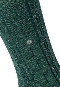 Calza di lana verde scuro caratterizzata da un design a trama strutturata e un dettaglio del logo in metallo argentato. La texture include fenditure più chiare distribuite in tutto.