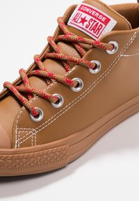 Zapatilla Converse de cuero marrón con cordones texturizados en rojo y marrón, ojales plateados y costuras blancas. Presenta una puntera de goma y una suela con patrón.