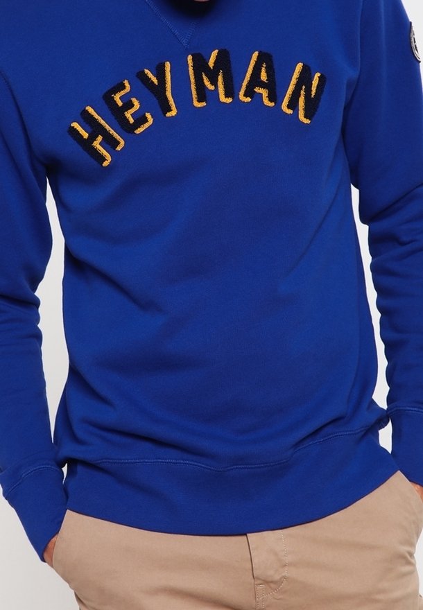 Sudadera azul con la inscripción "HEY MAN" bordada en el pecho en letras amarillas y negras. Material de algodón con puños acanalados.