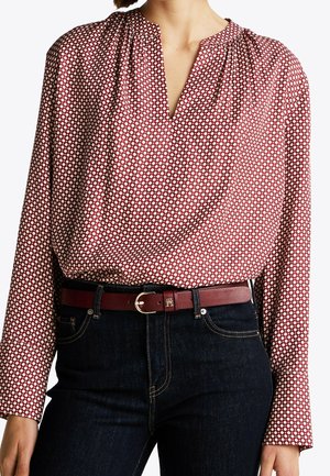 Femme portant un chemisier à manches longues rouge et blanc à motifs, rentré dans un jean foncé avec une ceinture en cuir rouge avec une boucle dorée.